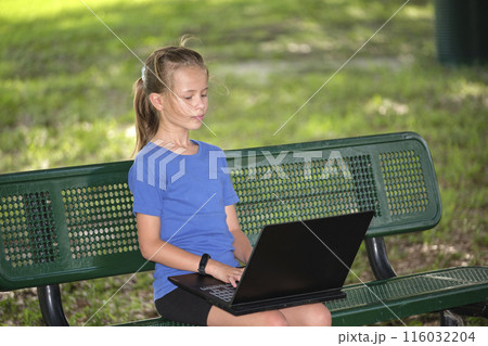 Cute teenager girl typing on laptop computer sitting on bench outdoors on summer sunny day. Education during quarantine concept Cute teenager girl typing on laptop computer sitting on bench outdoors on summer sunny day. Education during quarantine concept 116032204