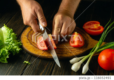 A chef hands use a knife on a cutting board to slice a fresh ripe tomato to prepare a vegetable breakfast dish 116032491