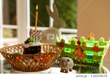 Easter cake with a lit candle. Easter colorful eggs in a basket on the table Easter cake with a lit candle. Easter colorful eggs in a basket on the table 116038676