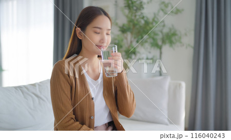 Happy young Asian woman drinking water while sitting on the sofa in the living room Happy young Asian woman drinking water while sitting on the sofa in the living room 116040243
