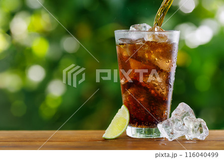 cola pouring in glass with ice cubes on wooden table outdoors 116040499