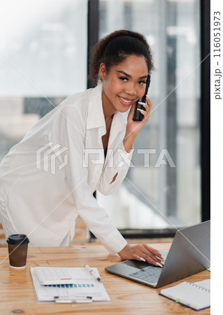 Confident young woman in white shirt multitasking with phone and laptop in a bright, modern office environment. Confident young woman in white shirt multitasking with phone and laptop in a bright, modern office environment. 116051973
