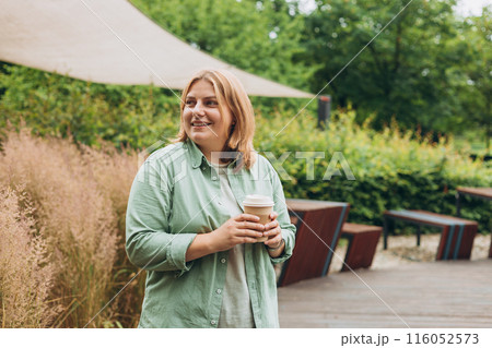 Happy redhead 30s Woman holding brown paper cup of coffee on public park background, Coffee to go on the city street. Food, rest, Take away concept. 116052573
