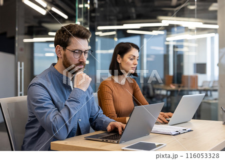 Two focused professionals working together at a desk with laptops in a contemporary office setting, conveying collaboration and teamwork. Two focused professionals working together at a desk with laptops in a contemporary office setting, conveying collaboration and teamwork. 116053328