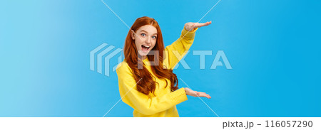Excited lively good-looking redhead ecstatic girl smiling fascinated and amused, showing big, large object, shaping size with raised hands over blue background, standing joyful, impressed 116057290