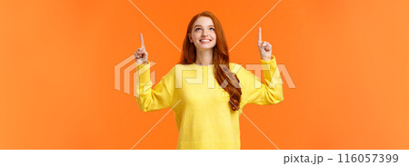 Dreamy happy and upbeat redhead young modern woman gazing up with satisfied smile. Gorgeous girl ginger hair conemplate top advertisement, pointing upwards, standing orange background 116057399