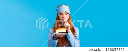 Indecisive redhead girl cant resist temptation, biting finger and looking with desire at slice cake, want eat dessert but worry calories, standing blue background in nightwear and sleep mask 116057532
