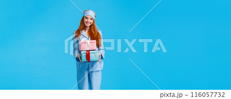Cheerful and lovely young happy redhead woman waking up early christmas eve congratulating with winter holidays, wearing pyjama and sleep mask, holding boxes with gifts, giving presents 116057732