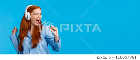 Sassy good-looking redhead female in pyjama having fun, listening music in big white headphones, singing into toothbrush as brushing teeth, getting ready go out, standing blue background Sassy good-looking redhead female in pyjama having fun, listening music in big white headphones, singing into toothbrush as brushing teeth, getting ready go out, standing blue background 116057761