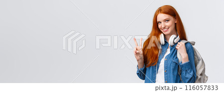 Lovely flirty and cheeky redhead college girl, wear headphones over neck and backpack, showing peace sign and smiling alluring camera, standing happy over white background enjoy student life 116057833