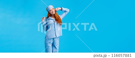 Full-length vertical shot cute and silly, teenage redhead girl, wearing lovely pyjama, waking up and stretching, had good sleep, smiling in morning, standing pleased blue background 116057881