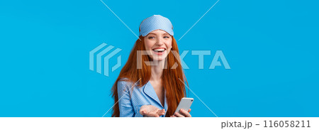 Happy and charismatic lovely redhead woman in nightwear, pyjama and sleep mask, laughing and saying what cute smiling, holding smartphone talking roommate, standing blue background 116058211