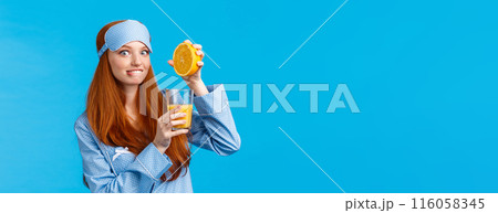Excited and cute pretty caucasian redhead woman in pyjama and sleep mask, biting lip tempting and eager try fresh made orange juice, squeeze fruit in glass and smiling camera, prepare drink 116058345