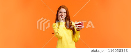 Happy lovely caucasian redhead female holding tasty cake with cream, touch cheek as receive congratulations being birthday girl, celebrating b-day with family, standing orange background Happy lovely caucasian redhead female holding tasty cake with cream, touch cheek as receive congratulations being birthday girl, celebrating b-day with family, standing orange background 116058558