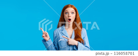 Surprised startled redhead girl found out special offer, discount promo, pointing upper right corner, wearing nightwear, gossiping about sleepover host, standing blue background 116058701