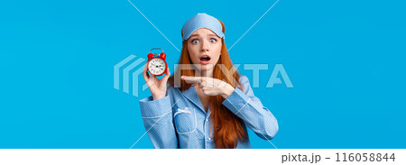 Shocked and concerned, worried cute redhead girl pointing at alarm clock with frustrated, nervous expression, being late, dont know how set up right time, standing blue background in nightwear Shocked and concerned, worried cute redhead girl pointing at alarm clock with frustrated, nervous expression, being late, dont know how set up right time, standing blue background in nightwear 116058844