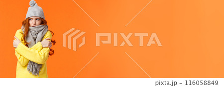 Its freezing cold outside. Cute redhead girl shaking and embracing herself to warm up, waiting someone on street in grey scarf, winter hat during snow and low temprature weather, orange background 116058849