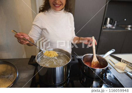 Close-up smiling woman taking freshly cooked farfalle pasta out of boiling water, preparing tomato sauce with seasonings and herbs, cooking a family dinner according to a traditional Italian recipe 116059948