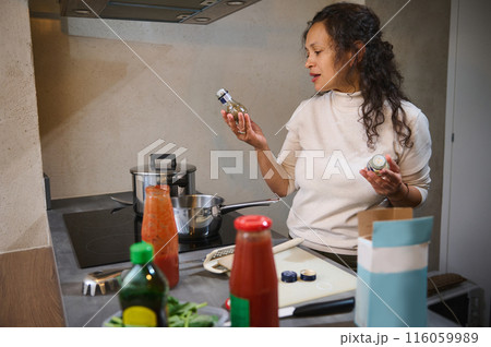 Beautiful curly hired brunette woman housewife holding small bottle with culinary herbs, standing at stove, cooking her favorite dish in minimalist modern kitchen interior. 116059989