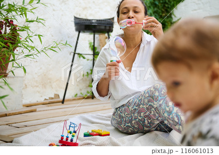 Cheerful woman in white shirt blowing bubbles while spending time with her little child in a lush green outdoor setting, evoking the joy and warmth of family time. 116061013