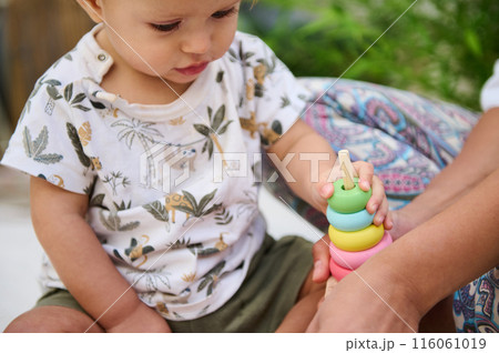 Selective focus on the hand of baby boy on colorful wooden circles of a pyramid, playing educational and developmental games, developing motor skills and thinking. Childhood and Kids entertainment 116061019