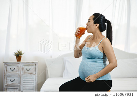 Pregnant woman in blue tank top drinking glass of orange juice, sitting indoors in softly lit bedroom Pregnant woman in blue tank top drinking glass of orange juice, sitting indoors in softly lit bedroom 116067931