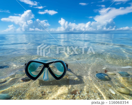 A snorkel mask rests on a clear, shallow beach with pebbles, under a bright blue sky with fluffy clouds. 116068354