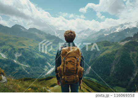 A solo traveler with a backpack stands on a mountain, overlooking a vast, green valley with winding paths, surrounded by majestic peaks under a cloudy sky 116069358
