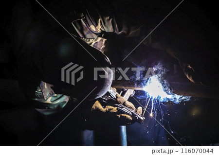 Welder at work. Man in a protective mask. The welder makes seams on the metal. Sparks and smoke when welding. Welder at work. Man in a protective mask. The welder makes seams on the metal. Sparks and smoke when welding. 116070044