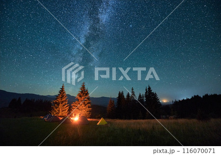 Night camping in mountains under starry sky. Tourist tents in campsite under sky full of stars with Milky way. Silhouette of hikers having a rest near burning campfire. Concept of tourism, adventures. Night camping in mountains under starry sky. Tourist tents in campsite under sky full of stars with Milky way. Silhouette of hikers having a rest near burning campfire. Concept of tourism, adventures. 116070071
