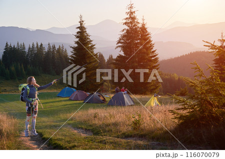 Woman traveller hiking in mountains at sunset. Young, sporty female hiking, carrying backpack, using trekking sticks, showing, pointing at sunset. Concept of tourism and adventure. 116070079