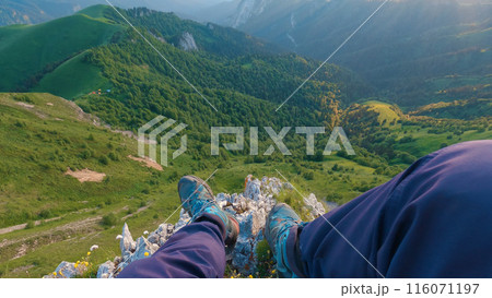 First-person view of legs of a man sitting on edge of a cliff overlooking a stunning mountainous landscape. The man is wearing blue pants and blue shoes, enjoying beauty of green hills and forests. 116071197