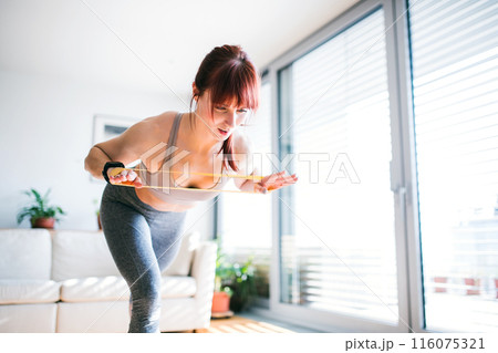Beautiful redhead woman's home fitness routine with resistance band. Home workout on gym mat in living room, sporty woman wearing sportswear. Beautiful redhead woman's home fitness routine with resistance band. Home workout on gym mat in living room, sporty woman wearing sportswear. 116075321