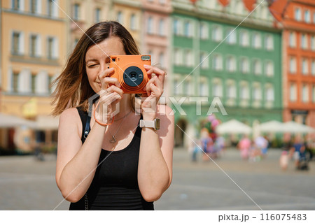 Woman taking photos in picturesque European city square, surrounded by colorful historic buildings. Female tourist captures memories during travel with vintage instant camera 116075483