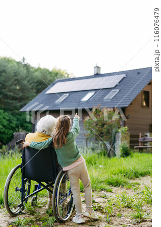 Granddaughter showing grandma solar panels on roof. Senior lady in wheelchair spending time in nature. Granddaughter showing grandma solar panels on roof. Senior lady in wheelchair spending time in nature. 116076479