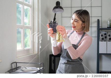 Housekeeper Cleaning and Organizing Home Office Space with a Smile, Wearing Apron and Gloves, Bright and Modern Interior 116080107