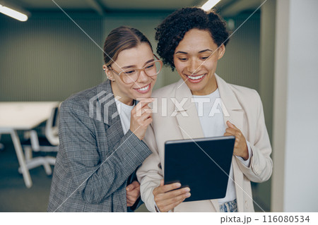 Cheerful female business people standing in modern coworking and using digital tablet together Cheerful female business people standing in modern coworking and using digital tablet together 116080534