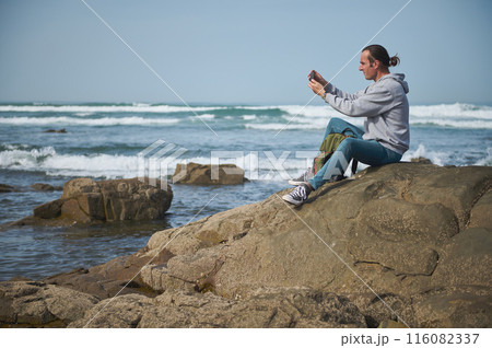 Man sitting on rocky shoreline taking photo with smartphone at the beach 116082337