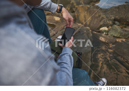 Person using smartphone outdoors while sitting on rocky terrain wearing casual attire Person using smartphone outdoors while sitting on rocky terrain wearing casual attire 116082372