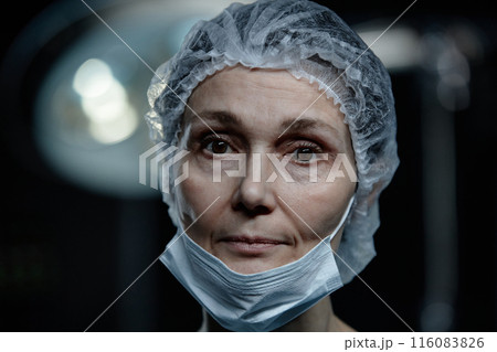 Close up portrait of mature woman as surgeon looking at camera standing in operating room set with dramatic lighting, copy space 116083826
