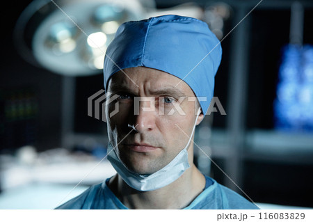 Close up portrait of tired male surgeon looking at camera standing in operating room with low light Close up portrait of tired male surgeon looking at camera standing in operating room with low light 116083829