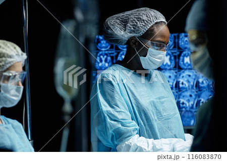 Side view portrait of African American woman as female surgeon wearing full protective clothing in operation with screens and monitors in background copy space 116083870