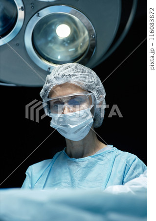 Vertical portrait of mature female surgeon wearing mask performing operation in blue light at hospital Vertical portrait of mature female surgeon wearing mask performing operation in blue light at hospital 116083872