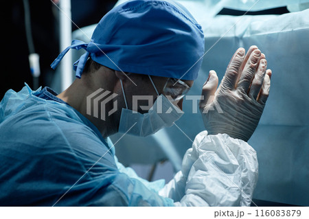 Side view portrait of male surgeon wearing mask and full protective clothing doing prayer ritual during surgery in operating room 116083879
