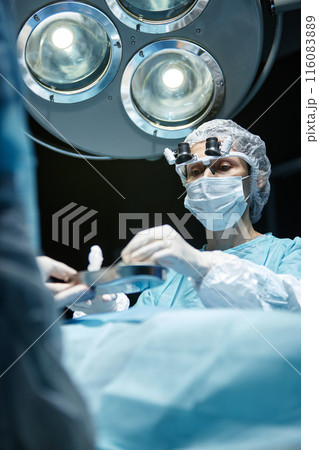 Vertical low angle portrait of mature female surgeon performing operation under surgical lamp in operating room 116083889
