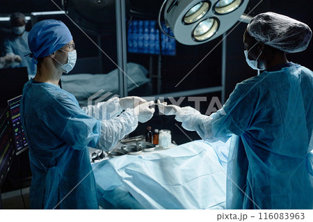 Side view portrait of medical assistant handing tools to surgeon performing surgery in operating room 116083963