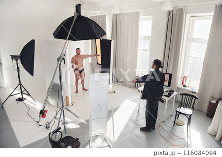 Wide angle view of male photographer directing model during professional photo shoot in white studio interior, copy space Wide angle view of male photographer directing model during professional photo shoot in white studio interior, copy space 116084094