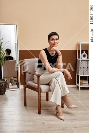 Vertical full length portrait of beautiful mature woman looking at camera sitting on chair posing in minimal interior Vertical full length portrait of beautiful mature woman looking at camera sitting on chair posing in minimal interior 116084360