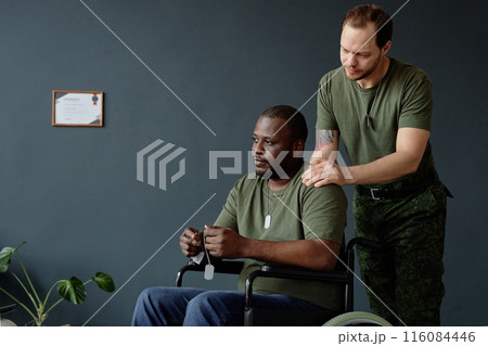 Minimal portrait of troubled Black man as army veteran with disability using wheelchair and looking away with friend soldier assisting copy space Minimal portrait of troubled Black man as army veteran with disability using wheelchair and looking away with friend soldier assisting copy space 116084446
