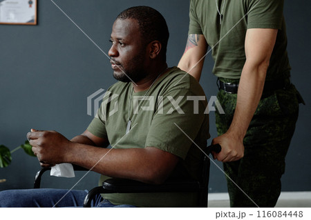 Side view portrait of grieving Black man as army veteran with disability using wheelchair and soldier assisting 116084448
